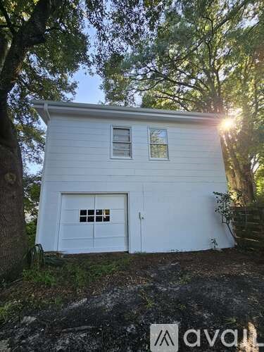 A small white building with a garage door is surrounded by trees.