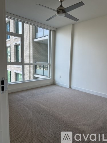 A room with a carpet floor, a ceiling fan, and a window overlooking a building.