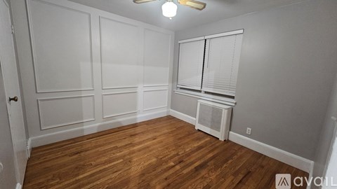 A room with wooden floors and white walls, featuring a ceiling fan and a window with blinds.