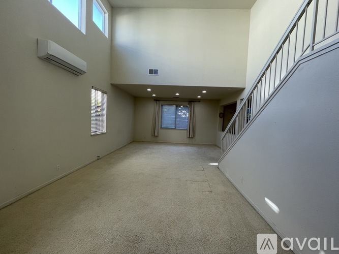A spacious room with a carpeted floor, a staircase with a railing, and a window with blinds.