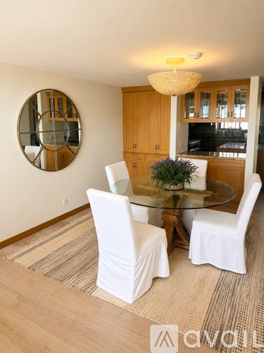 A round mirror hangs on a wall above a glass table with four white chairs around it.