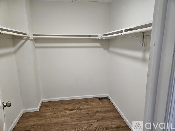 A room with white walls and wooden flooring, with a door on the left and a white shelf on the right.