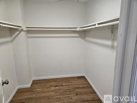 A room with white walls and wooden flooring, with a door on the left and a white shelf on the right.