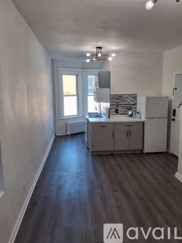 A room with wooden floors and white walls, with a kitchen area visible in the background.