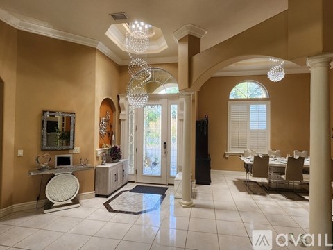 A well-lit, spacious living room with a chandelier, a dining table set for four, and a large window with blinds.