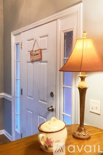 A lamp with a white shade sits on a table next to a door with a welcome sign hanging on it.