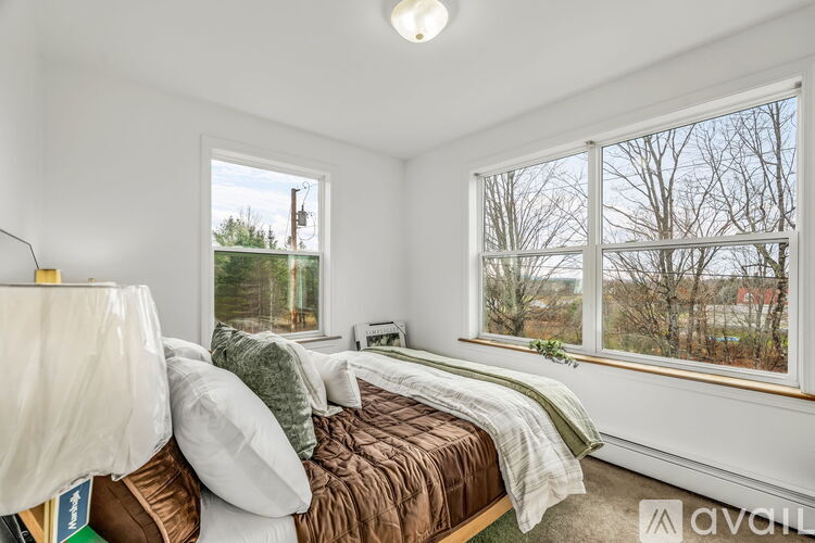 A bedroom with a bed covered in a brown comforter and pillows, a window with a view of trees and a building outside, and a lamp on the bedside table.