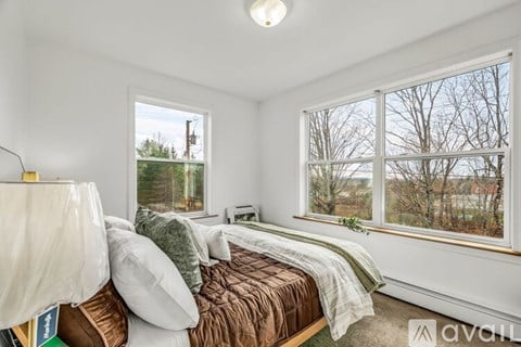 A bedroom with a bed covered in a brown comforter and pillows, a window with a view of trees and a building outside, and a lamp on the bedside table.