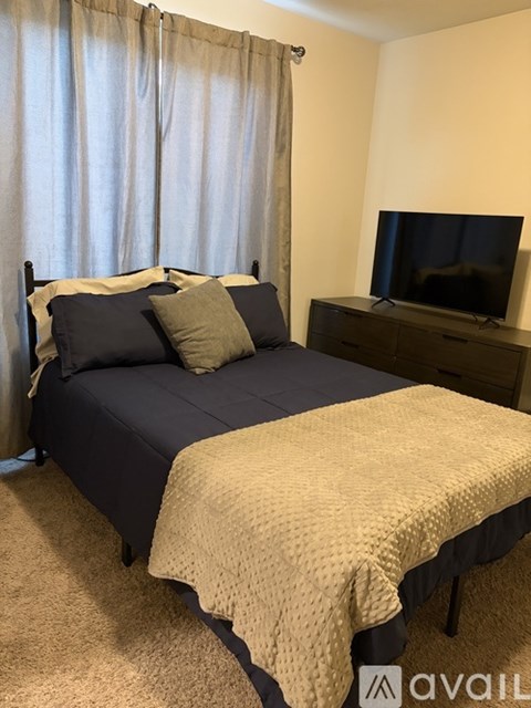 A bed with a blue comforter and a tan blanket is in the foreground of a room with a television in the background.