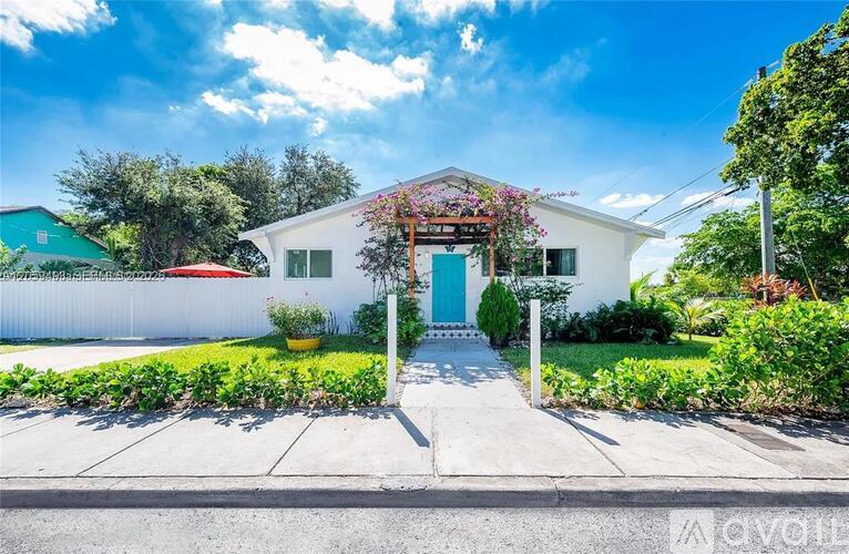 A white house with a blue door and a red awning is surrounded by greenery.