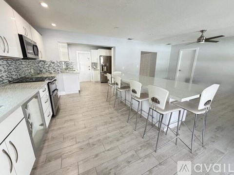 A kitchen with white cabinets and a grey countertop with a row of white chairs in front of it.