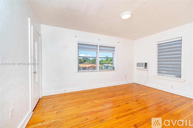 A room with wooden floors and white walls, featuring a window with a view of trees and a building outside.