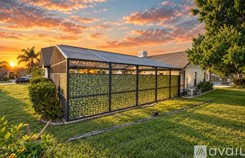A modern building with a glass wall and a metal roof is surrounded by a fence and greenery.