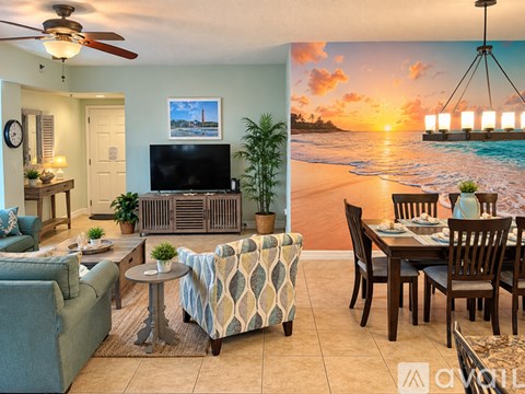 A living room with a television, a couch, a chair, a table, and a wall mural of a beach scene.
