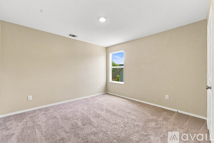A room with beige walls and carpeting, a window with a view of trees and a blue sky, and a door on the right.