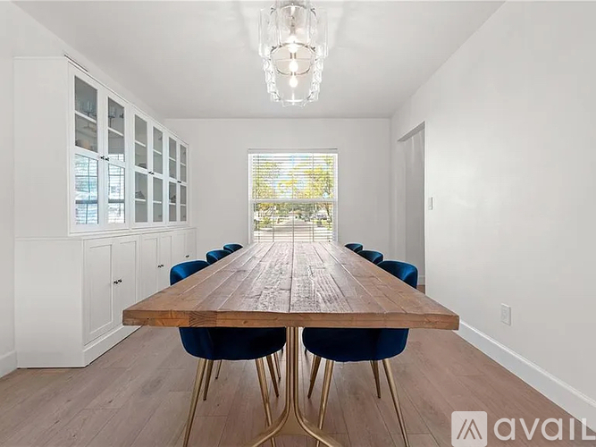 A long wooden table with blue chairs is in the middle of a room with white walls and a window.