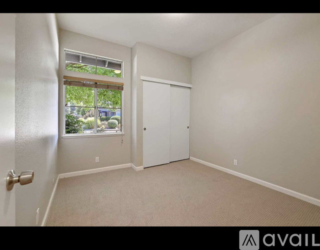 A room with a carpeted floor, a white door, and a window with a view of trees and a building outside.