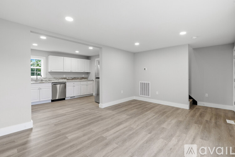 A spacious room with wooden flooring and white walls, leading to a kitchen area.