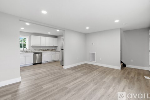 A spacious room with wooden flooring and white walls, leading to a kitchen area.
