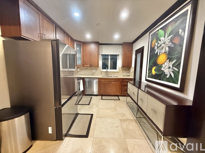 A kitchen with a refrigerator, trash can, and a painting of fruit on the wall.