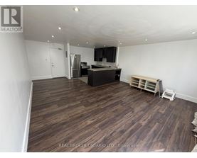 A room with wooden flooring and a kitchen area in the background.