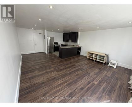 A room with wooden flooring and a kitchen area in the background.