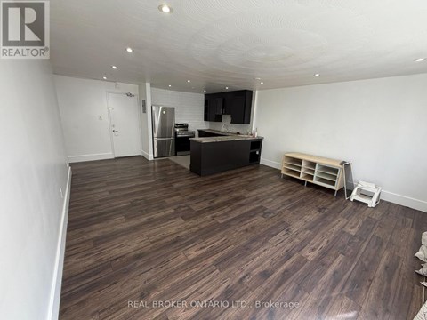 A room with wooden flooring and a kitchen area in the background.