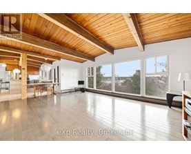 A spacious room with wooden ceiling and floor, furnished with a dining table and chairs, and a large window with a view of trees outside.