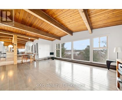 A spacious room with wooden ceiling and floor, furnished with a dining table and chairs, and a large window with a view of trees outside.
