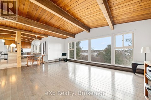 A spacious room with wooden ceiling and floor, furnished with a dining table and chairs, and a large window with a view of trees outside.