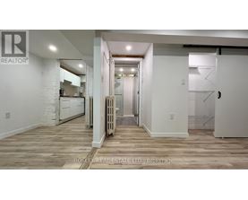 A spacious room with wooden flooring and white walls, with a kitchen area visible in the background.