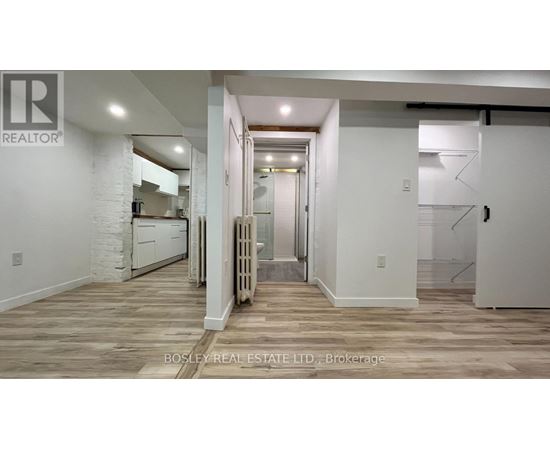 A spacious room with wooden flooring and white walls, with a kitchen area visible in the background.