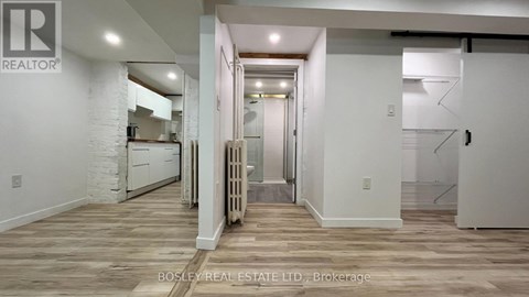 A spacious room with wooden flooring and white walls, with a kitchen area visible in the background.