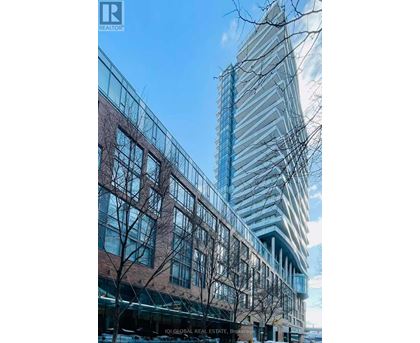 A tall glass building stands next to a shorter brick building with trees in front.