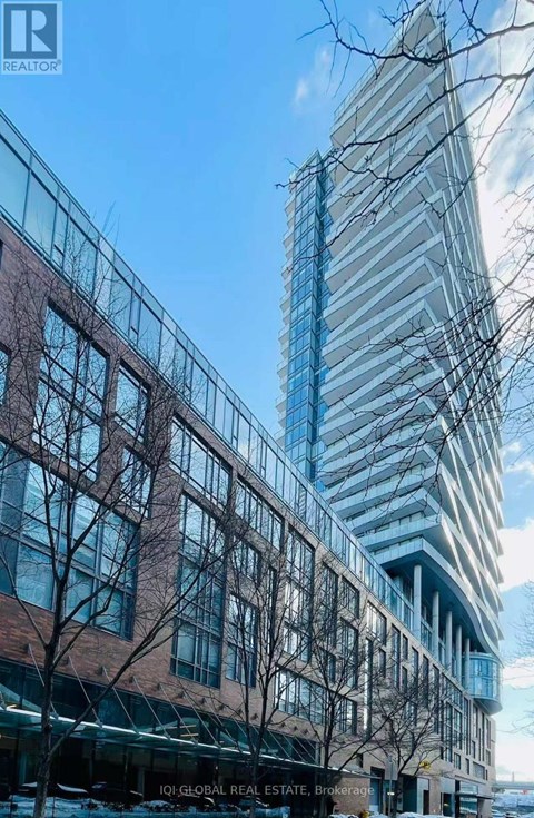 A tall glass building stands next to a shorter brick building with trees in front.