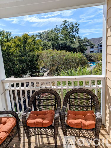 Two wicker chairs with orange cushions are on a balcony with a view of trees and a pool.