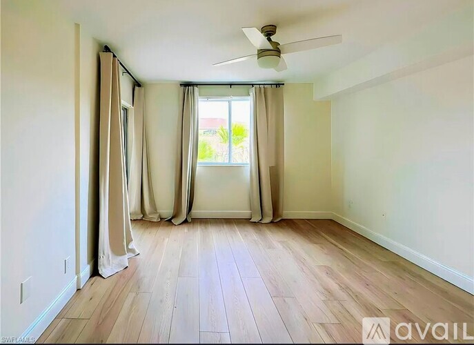 A room with a ceiling fan and light, wooden floors, and a window with curtains.
