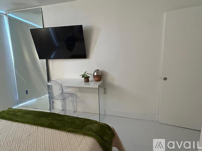 A black screen is mounted on a white wall above a glass desk with a chair and a small plant.