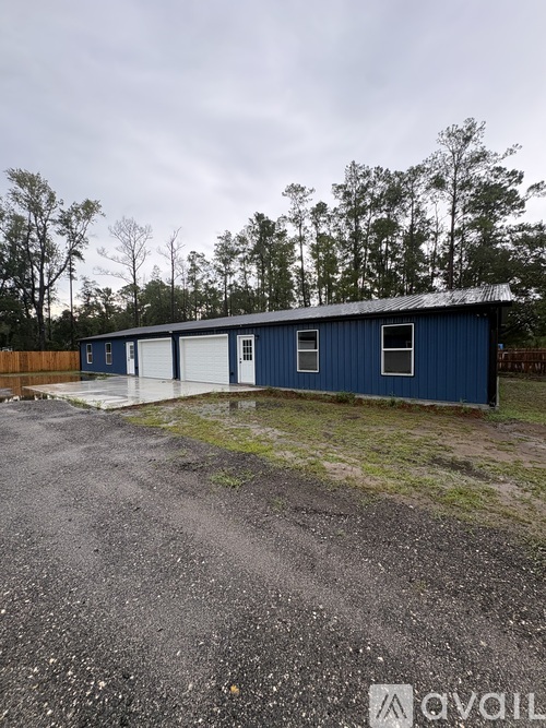 A blue building with a white door is surrounded by a gravel lot and trees.