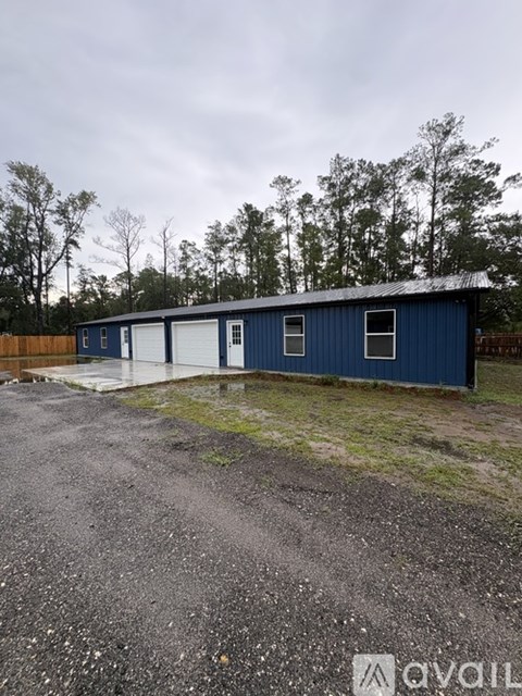 A blue building with a white door is surrounded by a gravel lot and trees.