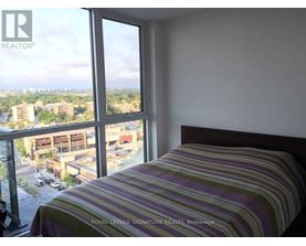 A bedroom with a striped bedspread and a large window overlooking a parking lot and buildings.