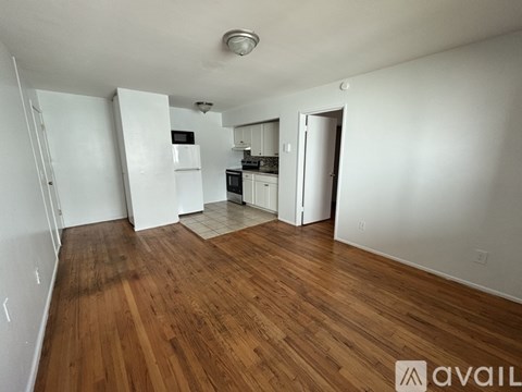 A room with wooden floors and white walls, with a kitchenette in the background.