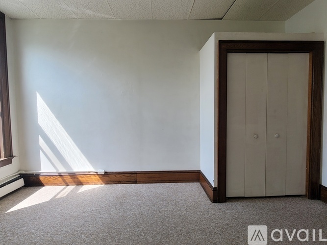 A room with a carpeted floor, a wooden door, and a window letting in sunlight.
