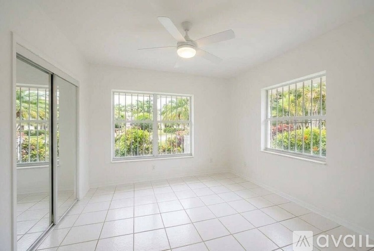 A room with a ceiling fan and two windows overlooking a green area.