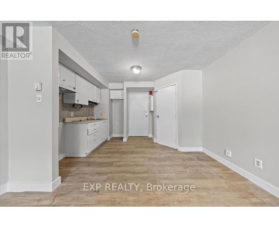 A white room with a kitchen area and a brokerage sign.