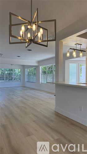 A modern chandelier hangs over a spacious room with wood flooring and large windows.