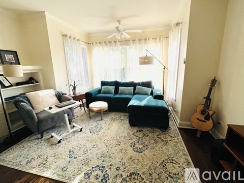 A living room with a grey couch, a blue ottoman, a rug, a chair, a table, a guitar, and a shelf with a lamp and a picture frame.