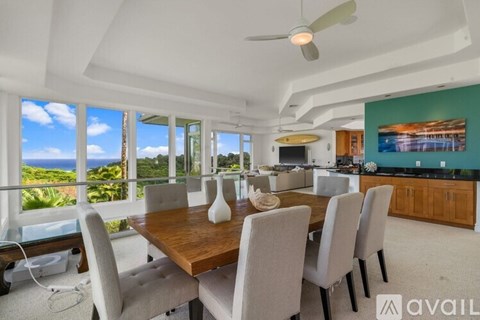 A dining room with a wooden table and chairs, a ceiling fan, and a large window overlooking the ocean.