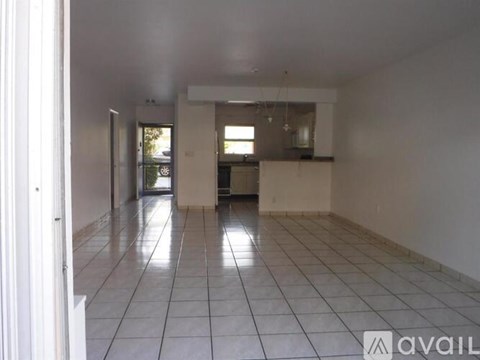 A spacious room with white tiled flooring and a kitchen area in the background.