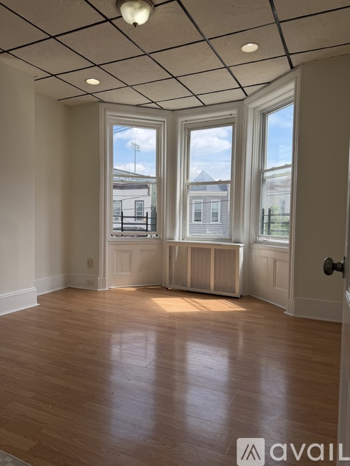 A room with wooden floors and white walls, with a window showing a view of a building and a balcony.